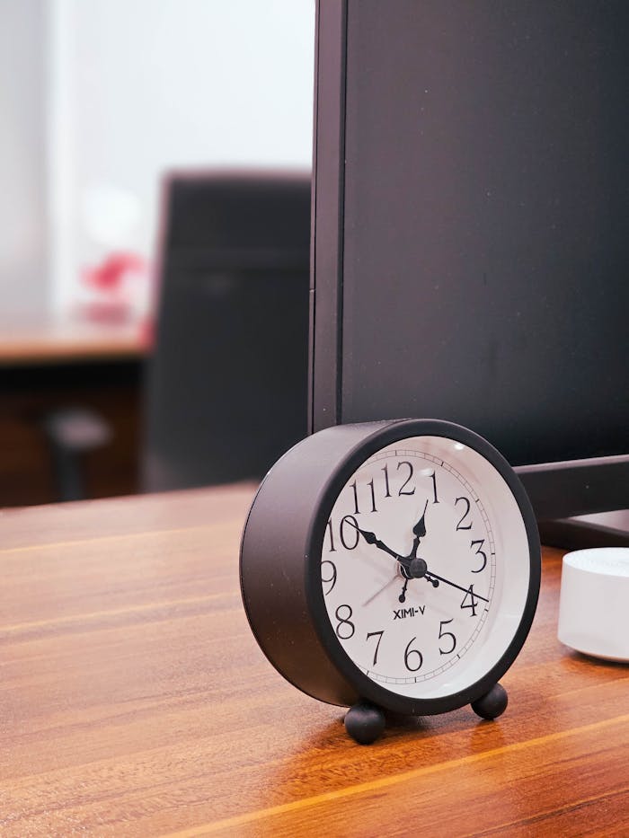 Black round clock on a wooden desk beside a computer monitor in an office setting.